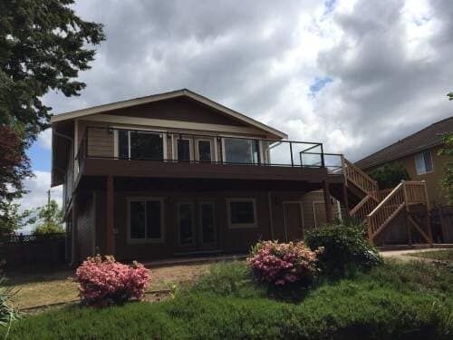 Two-story house with dark siding and a deck, surrounded by greenery and bushes. Cloudy sky in the background.