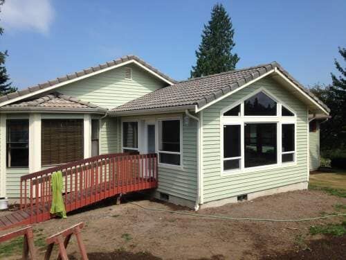 Exterior of a light green house with a wooden deck and large windows under a blue sky.