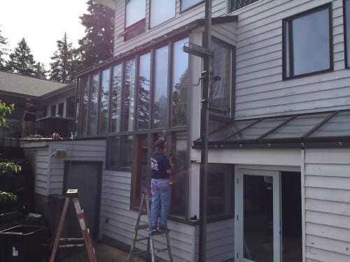 Person on ladder working on a building with large windows. Grey siding, black trim. Daylight.