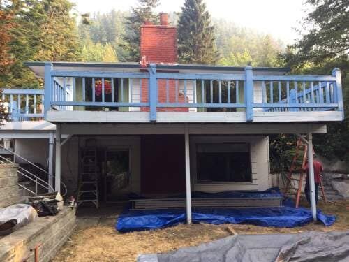 House with blue deck railing and red door, under construction, with a backdrop of trees.