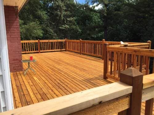 Wooden deck with stained railings and flooring, extending from a red brick wall, surrounded by trees.