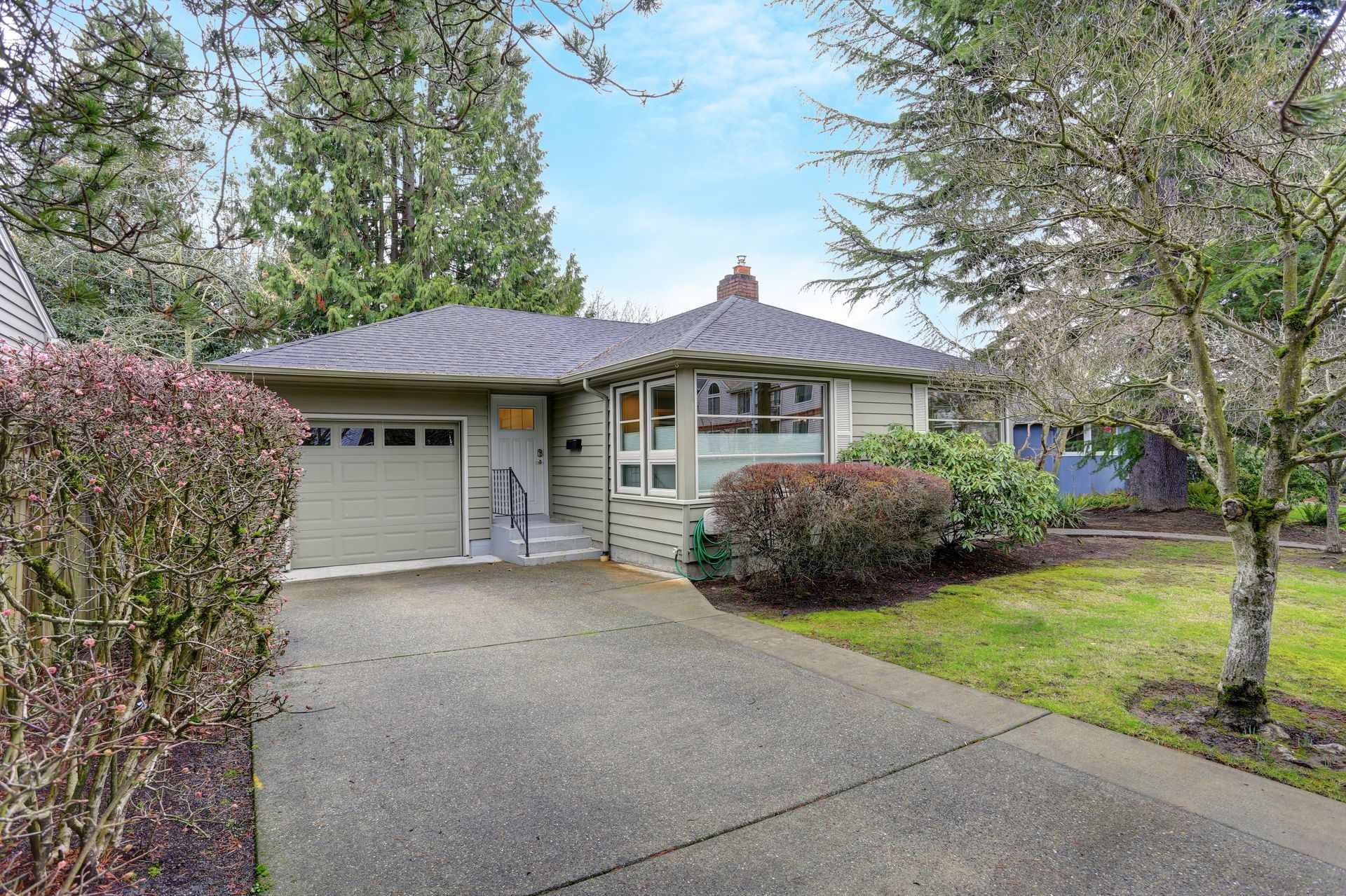 Green ranch-style house with gray driveway and garage, surrounded by bushes and trees.