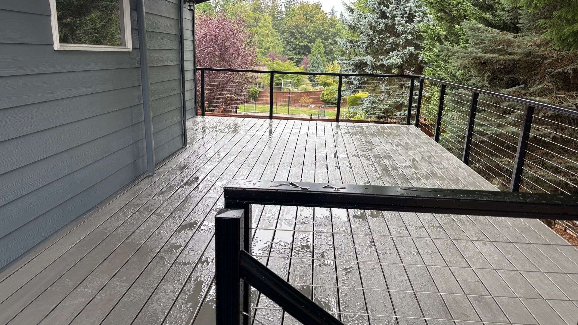 Wet wooden deck with black railing and view of trees in the rain.