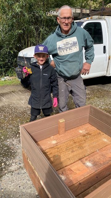 Man and child standing near a wooden box, in front of a white truck. Child holds a toy.