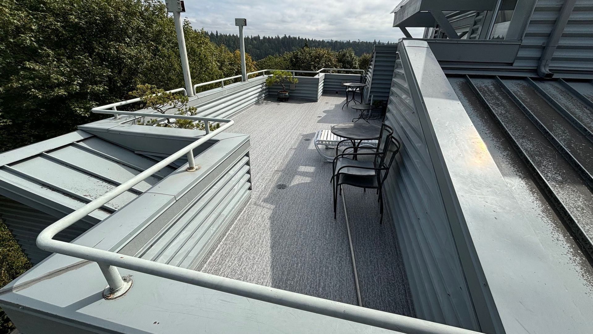 Rooftop patio with gray corrugated metal walls and roof, chairs, and greenery. Overcast sky.