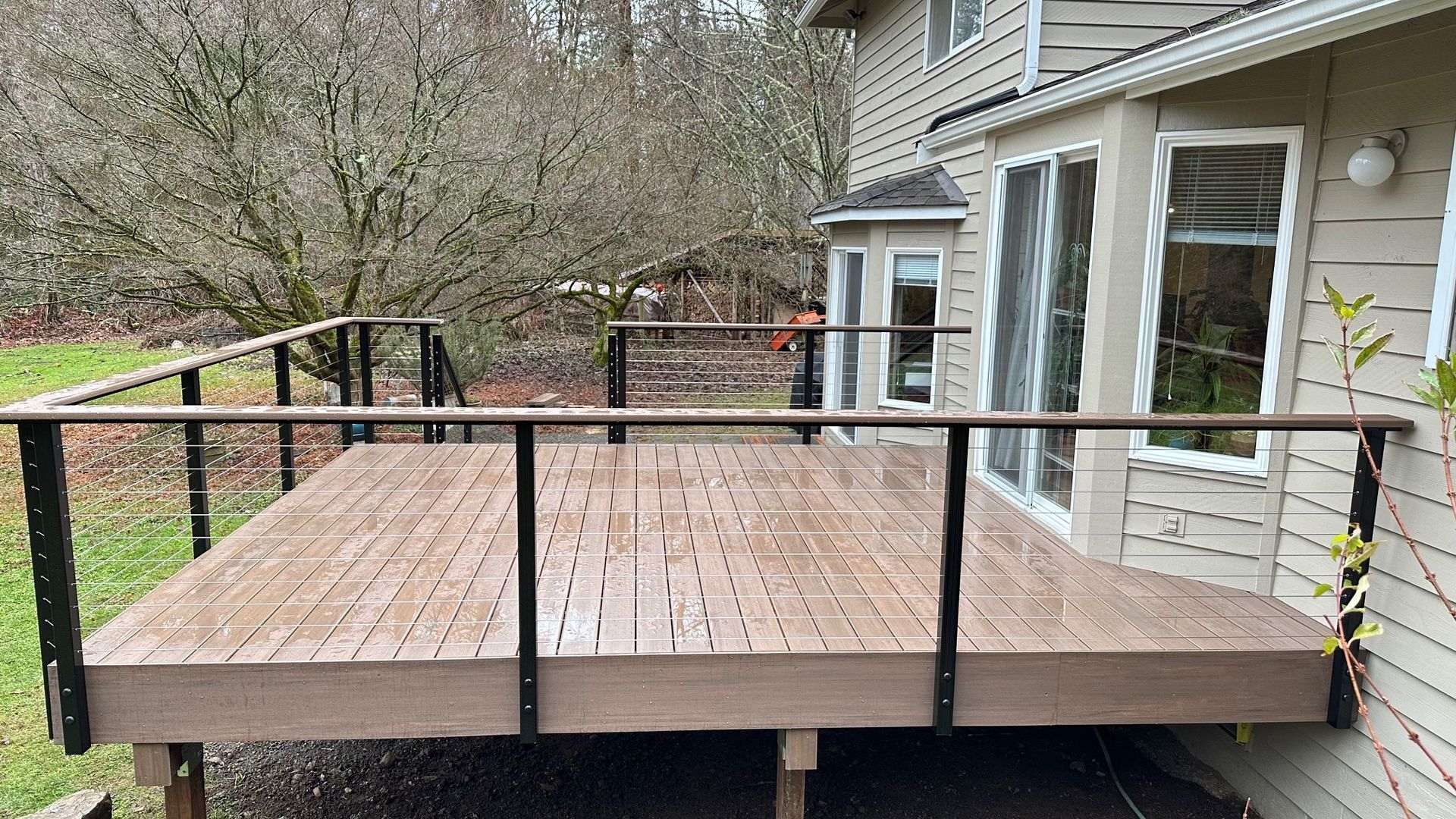 Wooden deck with black railing and cable infill, adjacent to a beige house, overlooking a yard with bare trees.