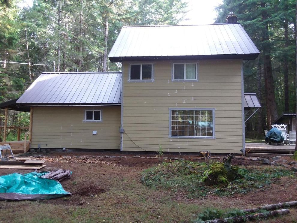 Two-story yellow house with metal roof in a wooded area. Porch and chimney are visible.