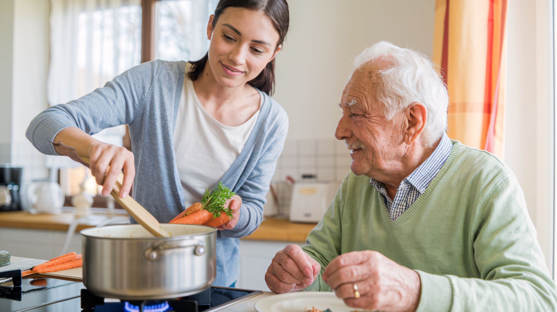 Woman stirring pot on stove while elderly man smiles at her in a kitchen.