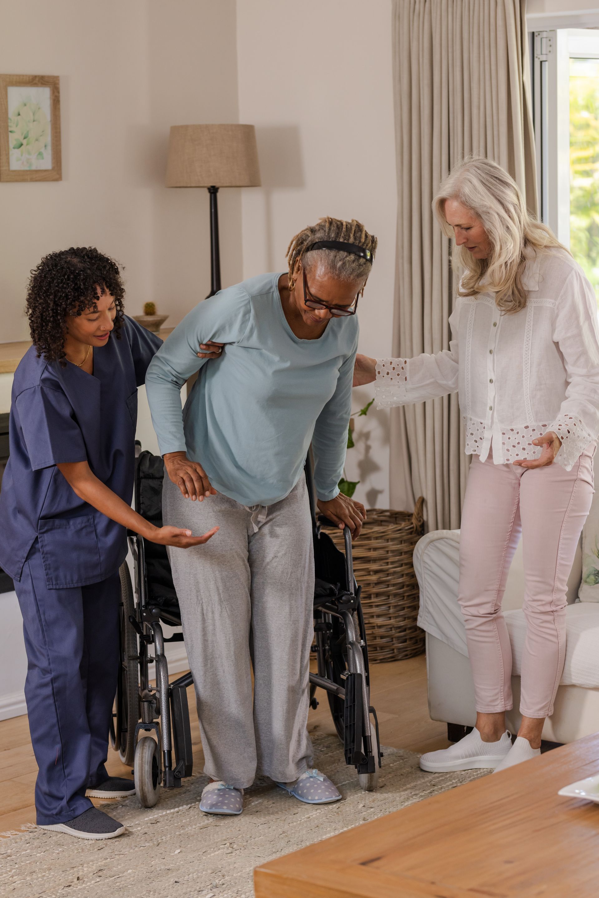 Two women helping an elderly woman stand from wheelchair. Inside a home.