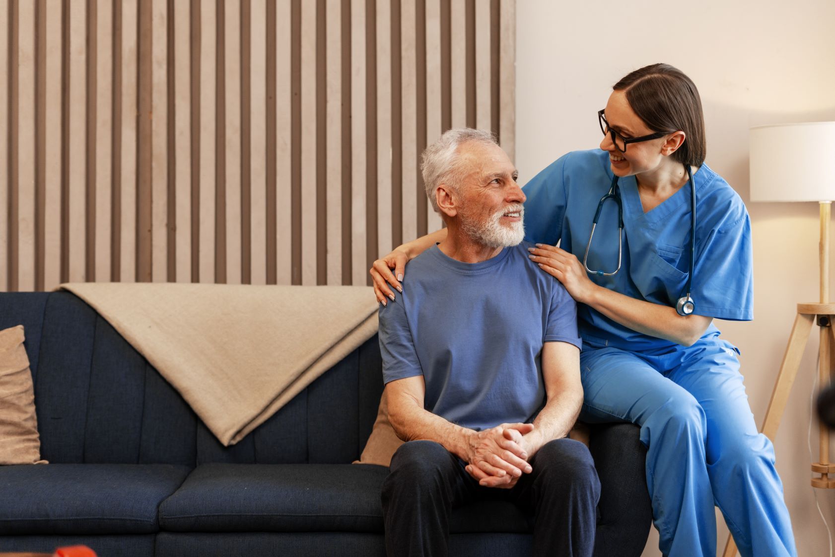 Nurse in blue scrubs comforts elderly man on a couch in a living room.