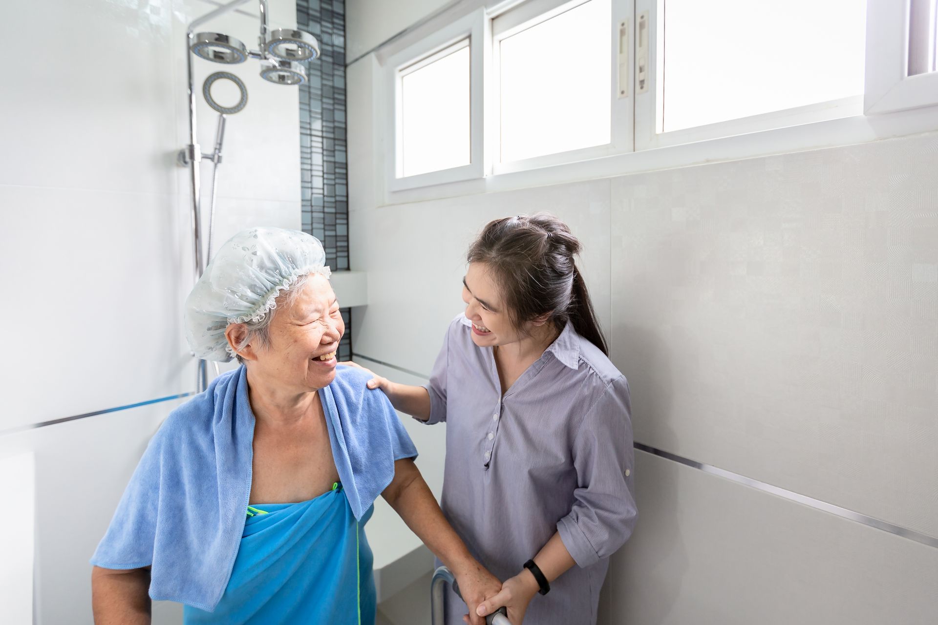 Woman helping elderly woman in a shower. Both smiling. Bathroom with window.