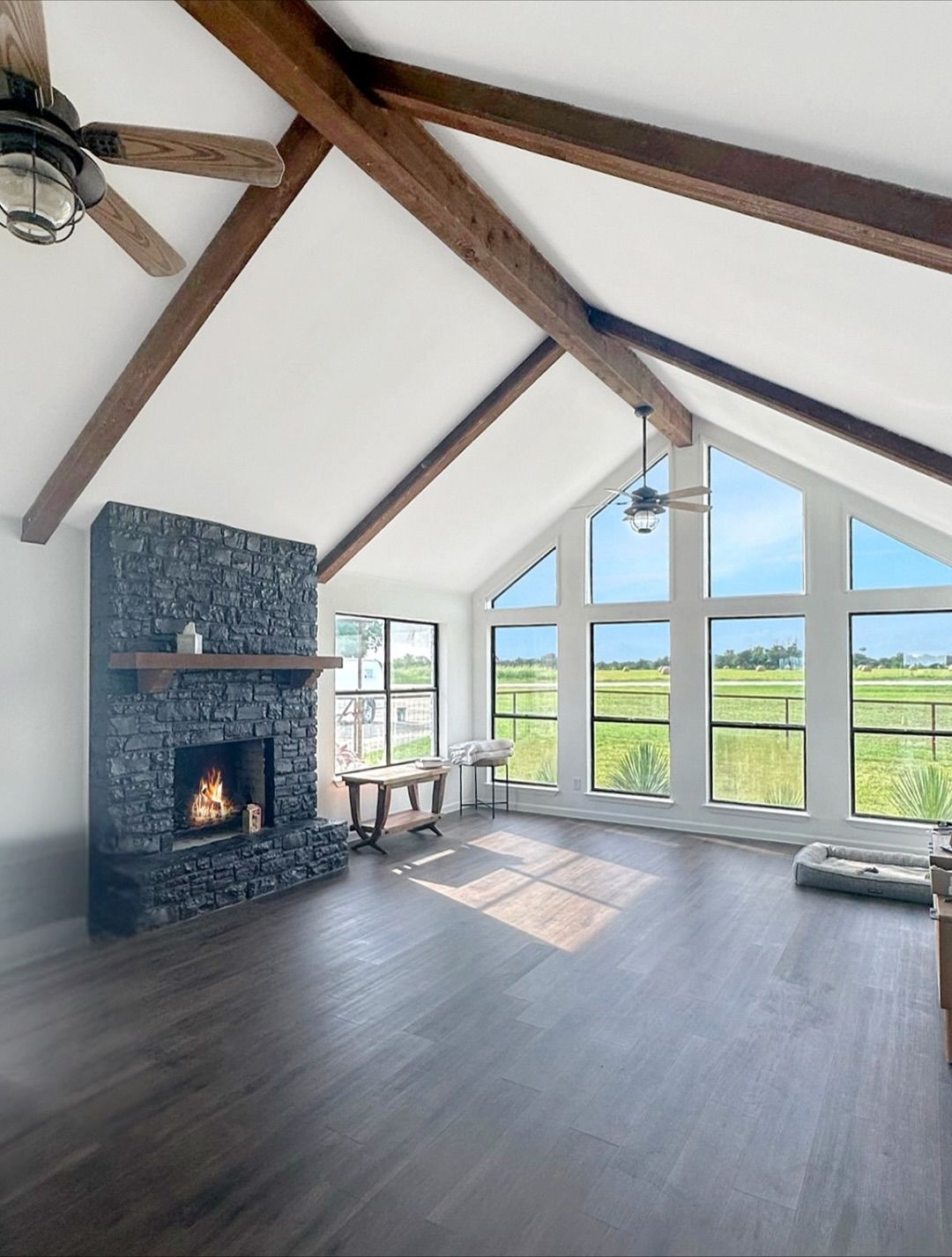 Living room with a stone fireplace, large windows, wood beams, and dark flooring.