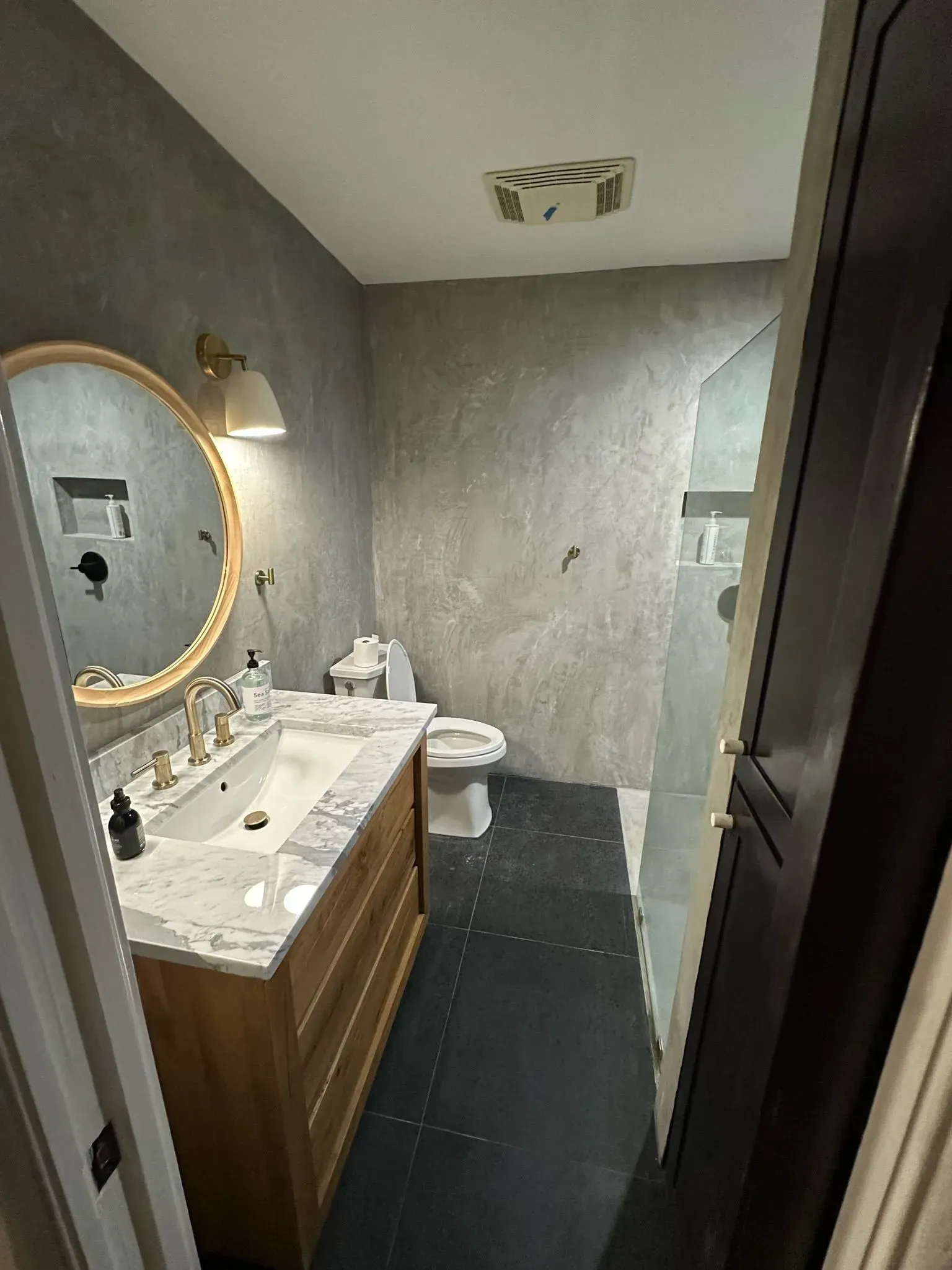 Bathroom with wood vanity, round mirror, gray walls, black floor, and shower.