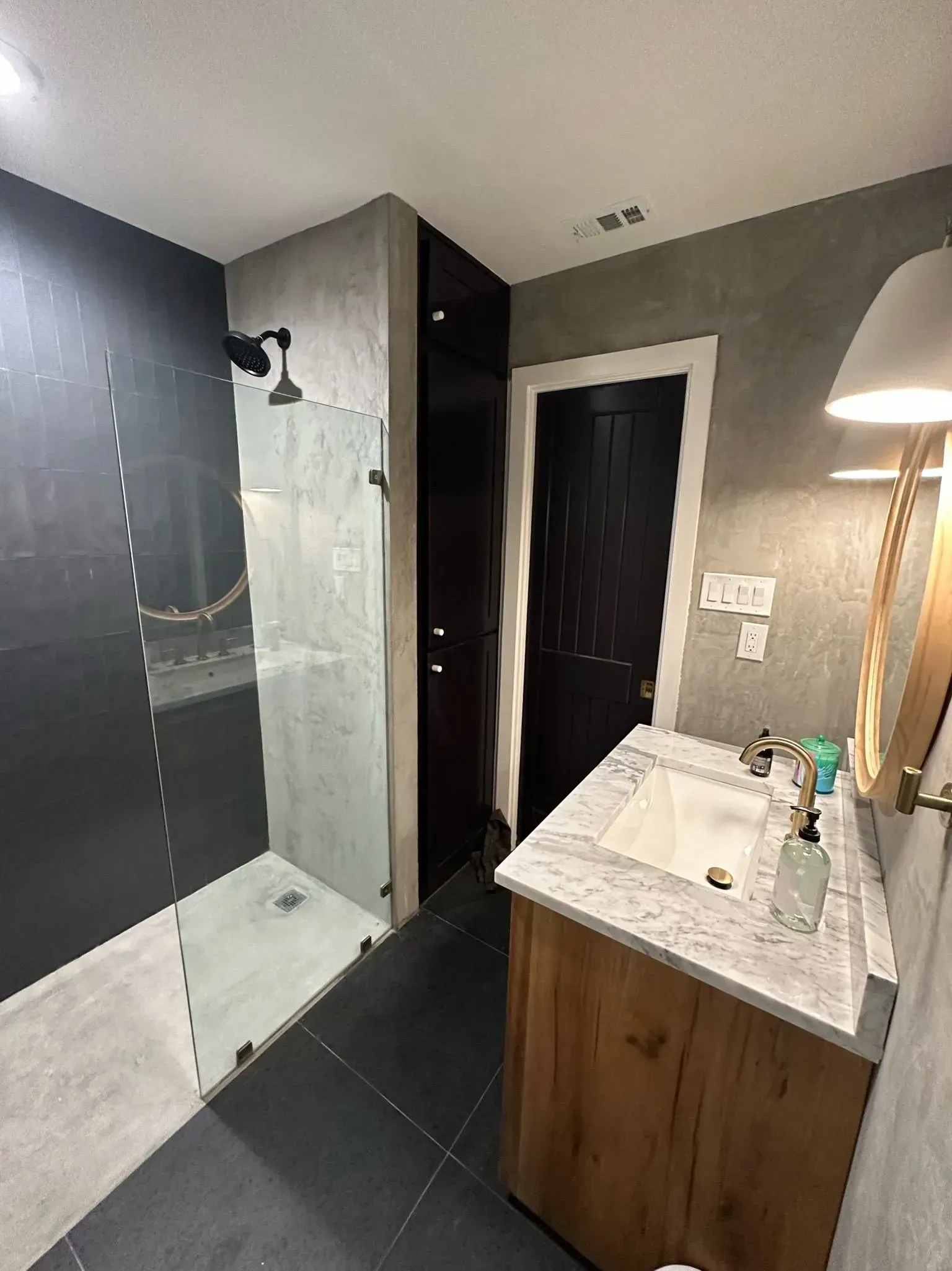 Bathroom with black tile, gray walls, glass shower, wooden vanity, and dark door.