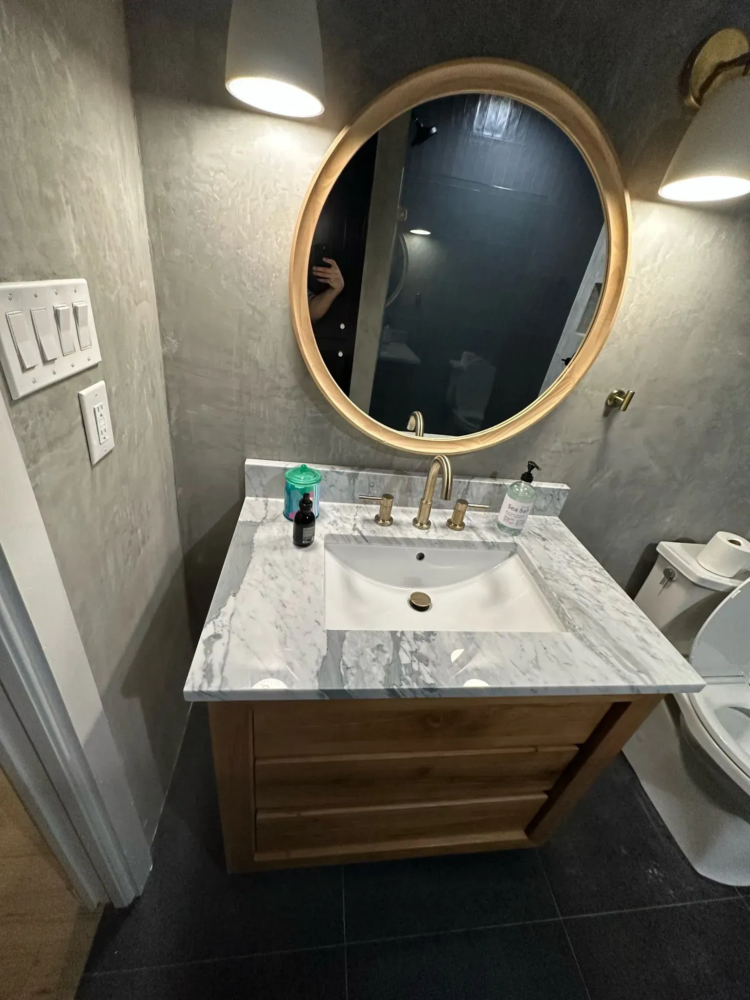 Bathroom with wooden vanity, marble countertop, round mirror, and gray walls.