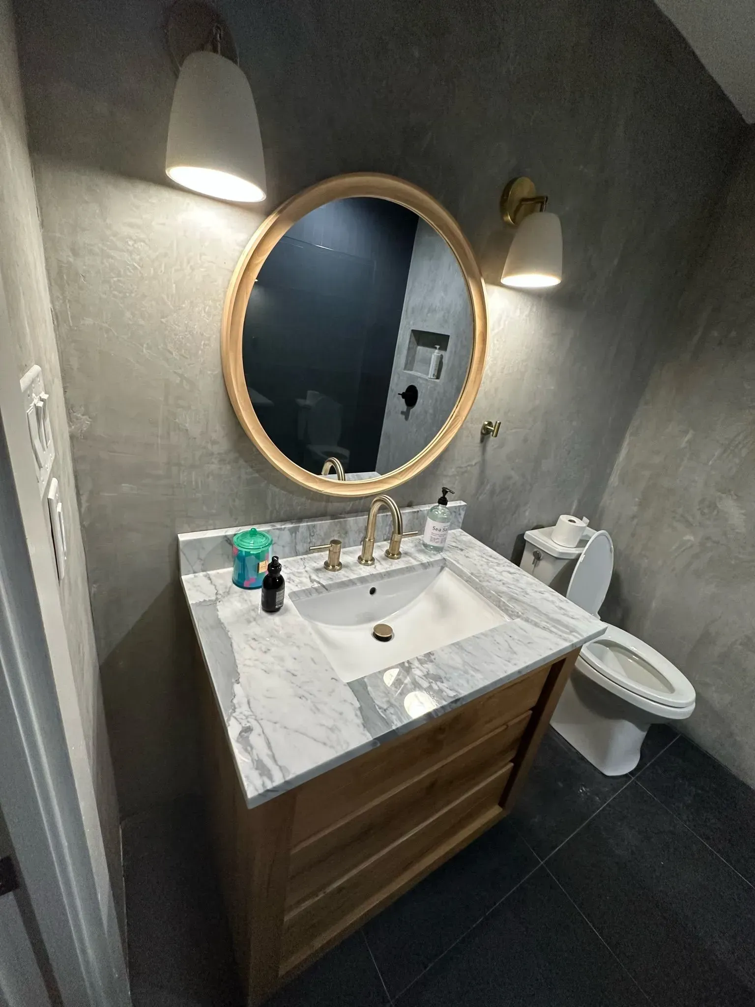 Bathroom with a wooden vanity, round mirror, and two sconces. Gray walls, white sink, and black tiled floor.