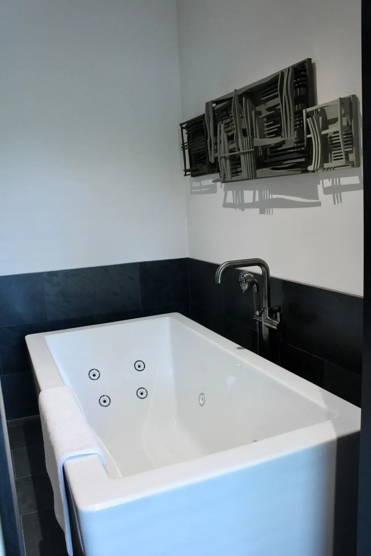 White jacuzzi bathtub with jets, against a dark tile wall. Chrome faucet and abstract wall art.