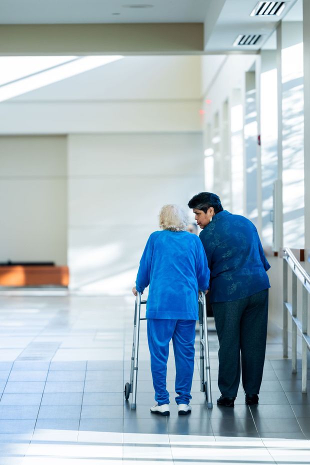 Two people walking in a bright hallway, one using a walker and the other supporting them.