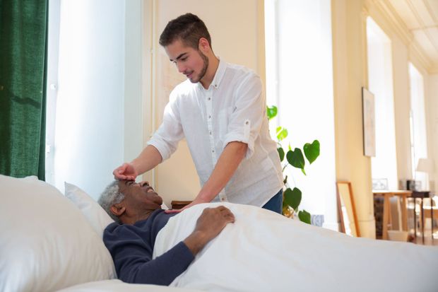 Caregiver checks a resting patient’s forehead in a bright bedroom or hospital room