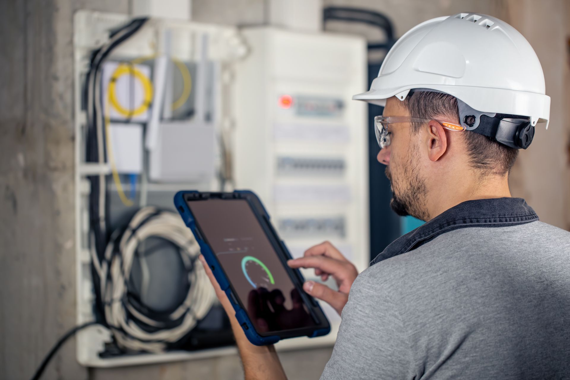 Technician in a white hard hat using a tablet to inspect electrical equipment in a utility room
