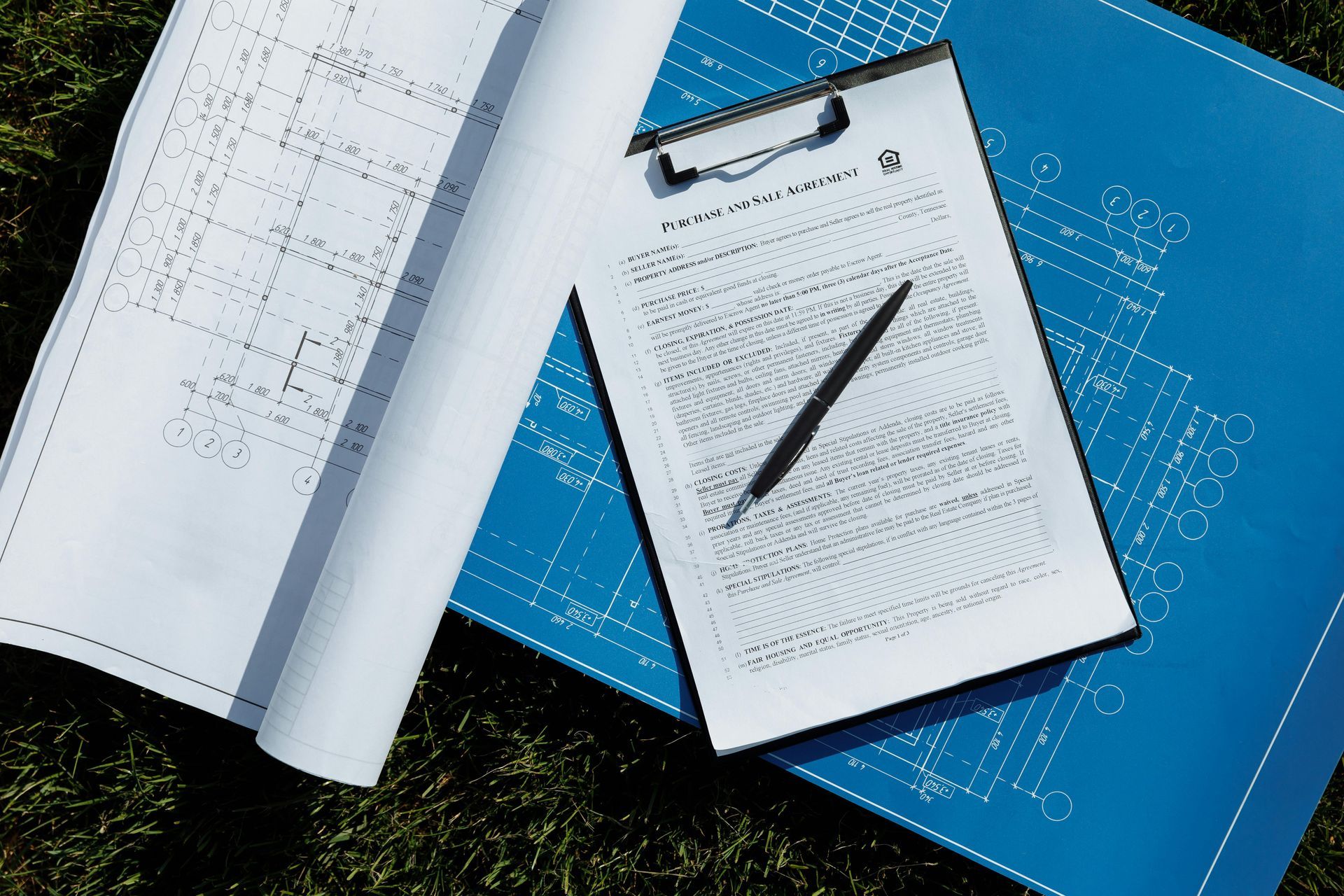 Clipboard with papers and pen beside rolled blueprints on grass