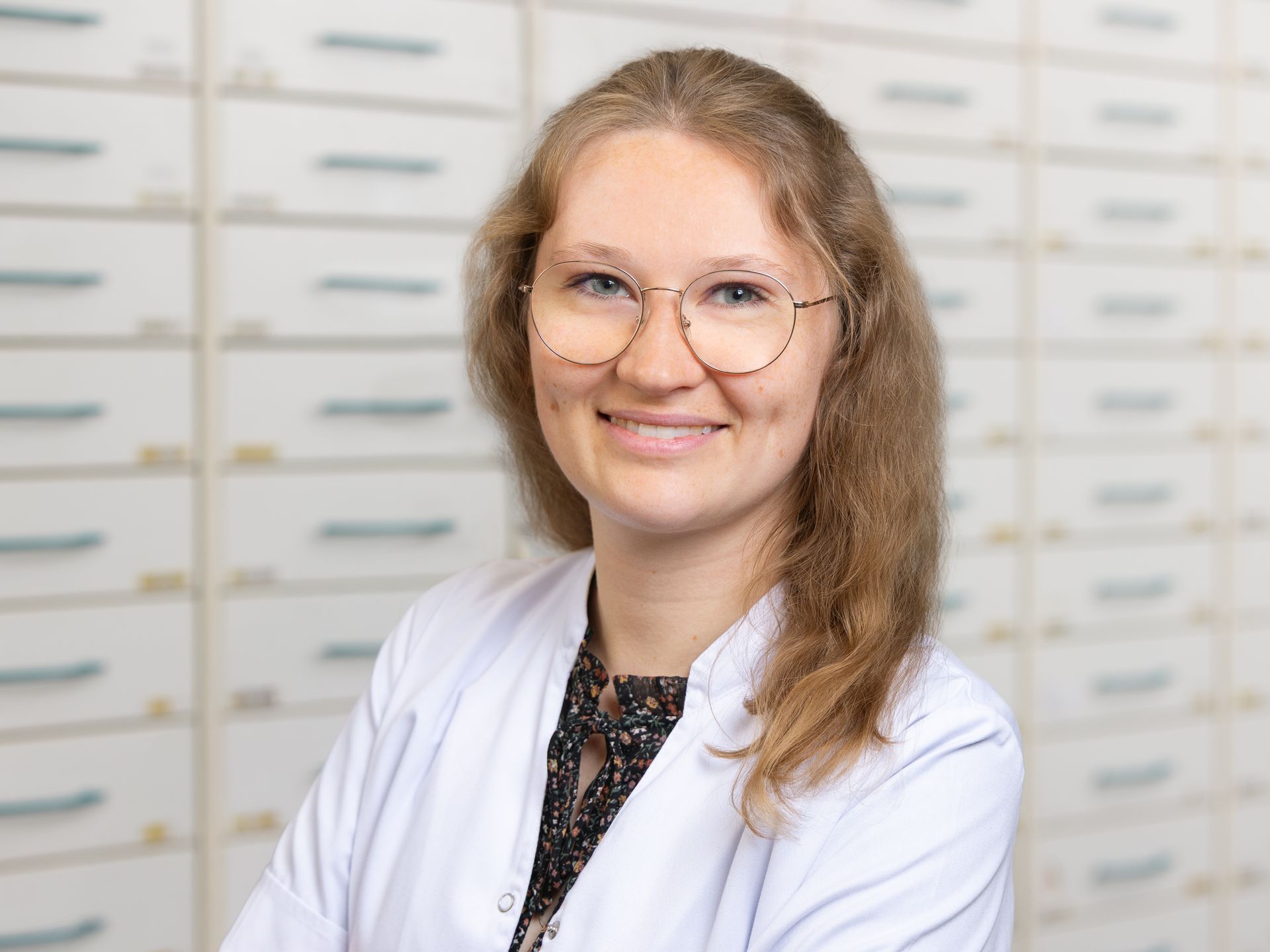 Eine Frau in einem weißen Laborkittel und mit Brille lächelt vor einer Apotheke.