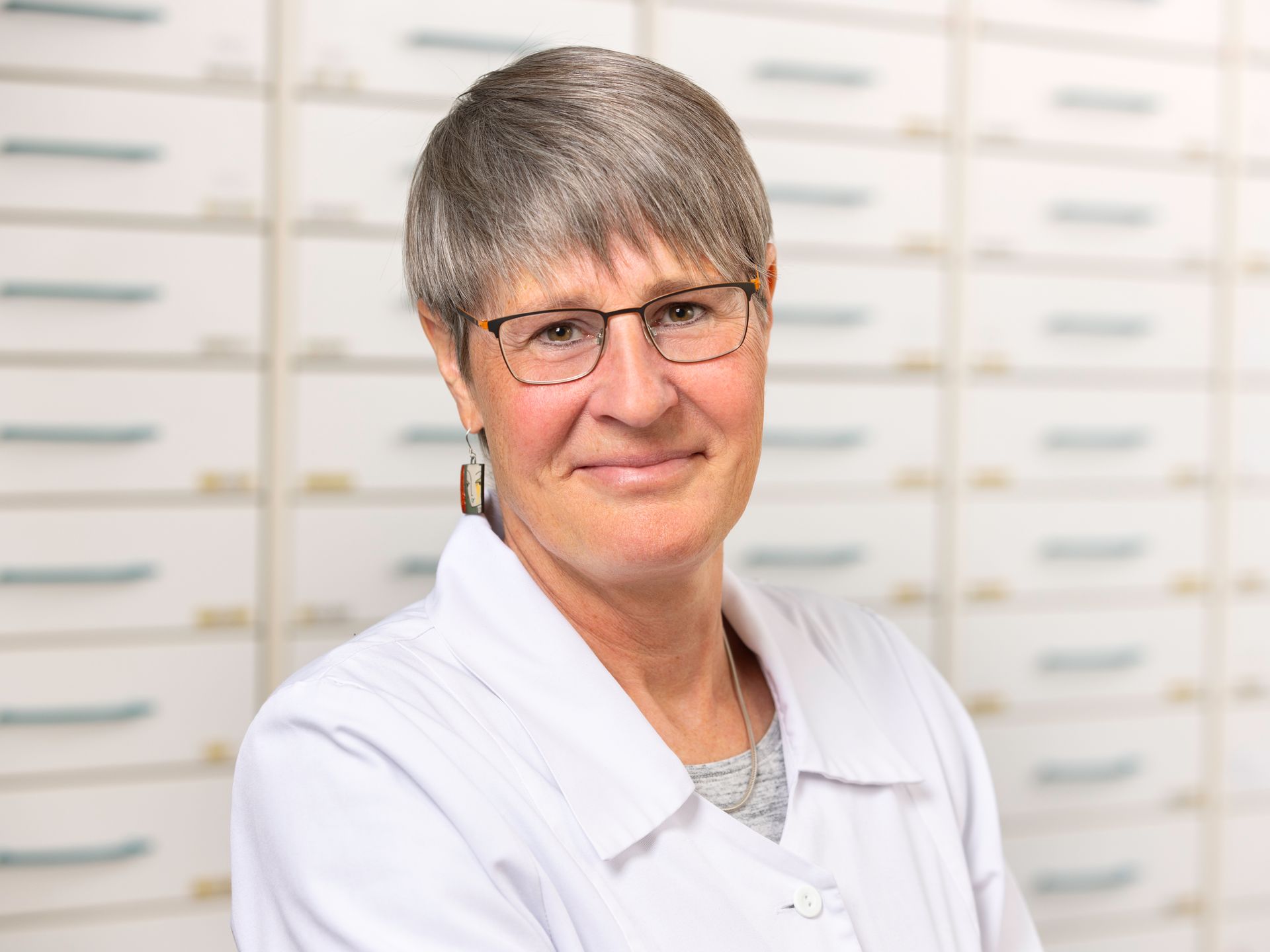 Eine Frau mit Brille und weißem Kittel steht vor dem Tresen einer Apotheke.