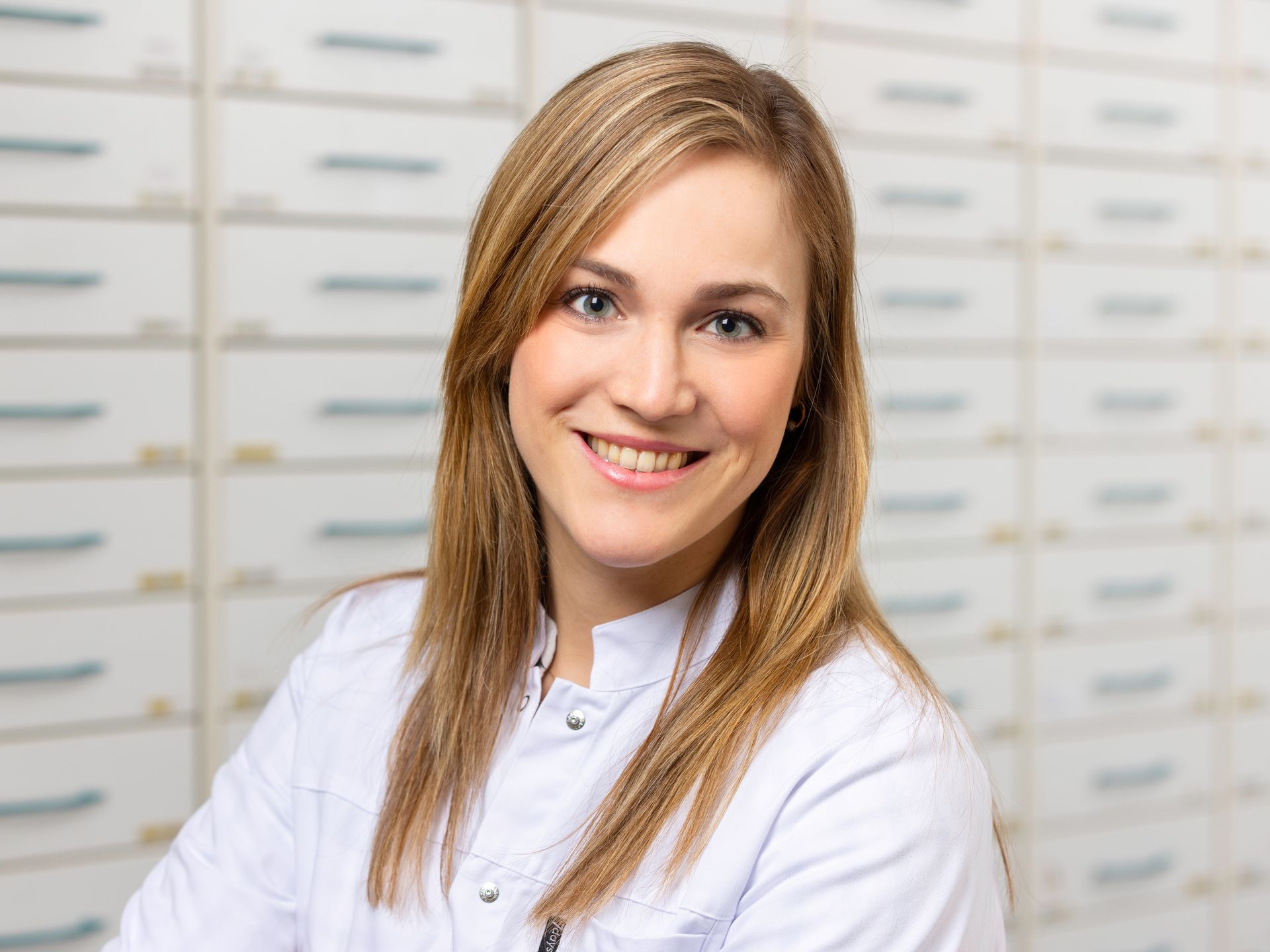 Eine Frau in einem weißen Laborkittel lächelt vor einer Apotheke.