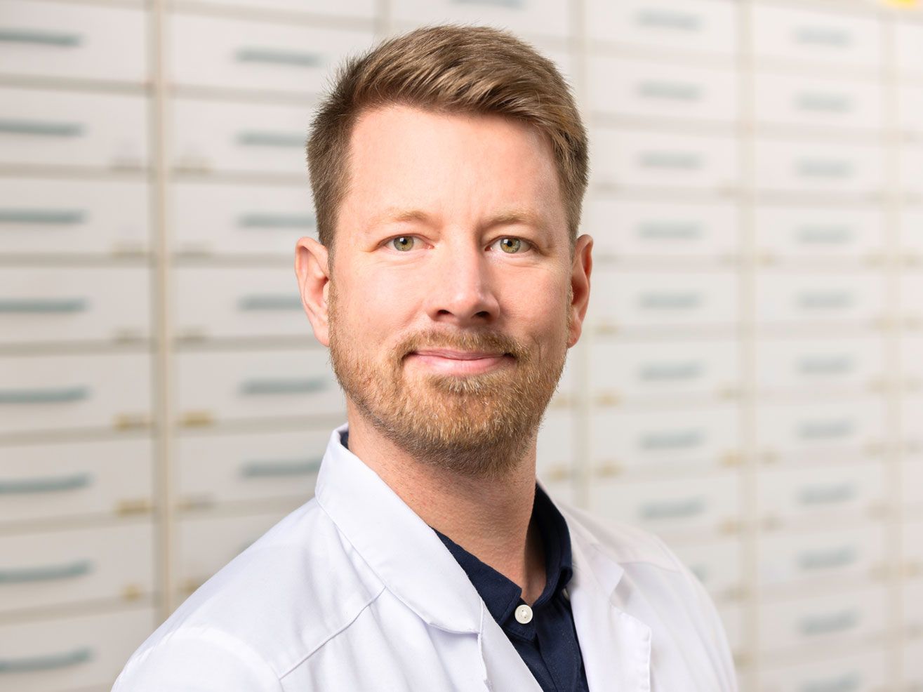 Ein Mann mit Bart trägt einen weißen Laborkittel und lächelt.