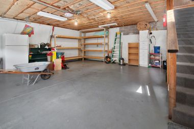 Empty garage with concrete floor, exposed wood ceiling, shelves, ladder, and stored household items