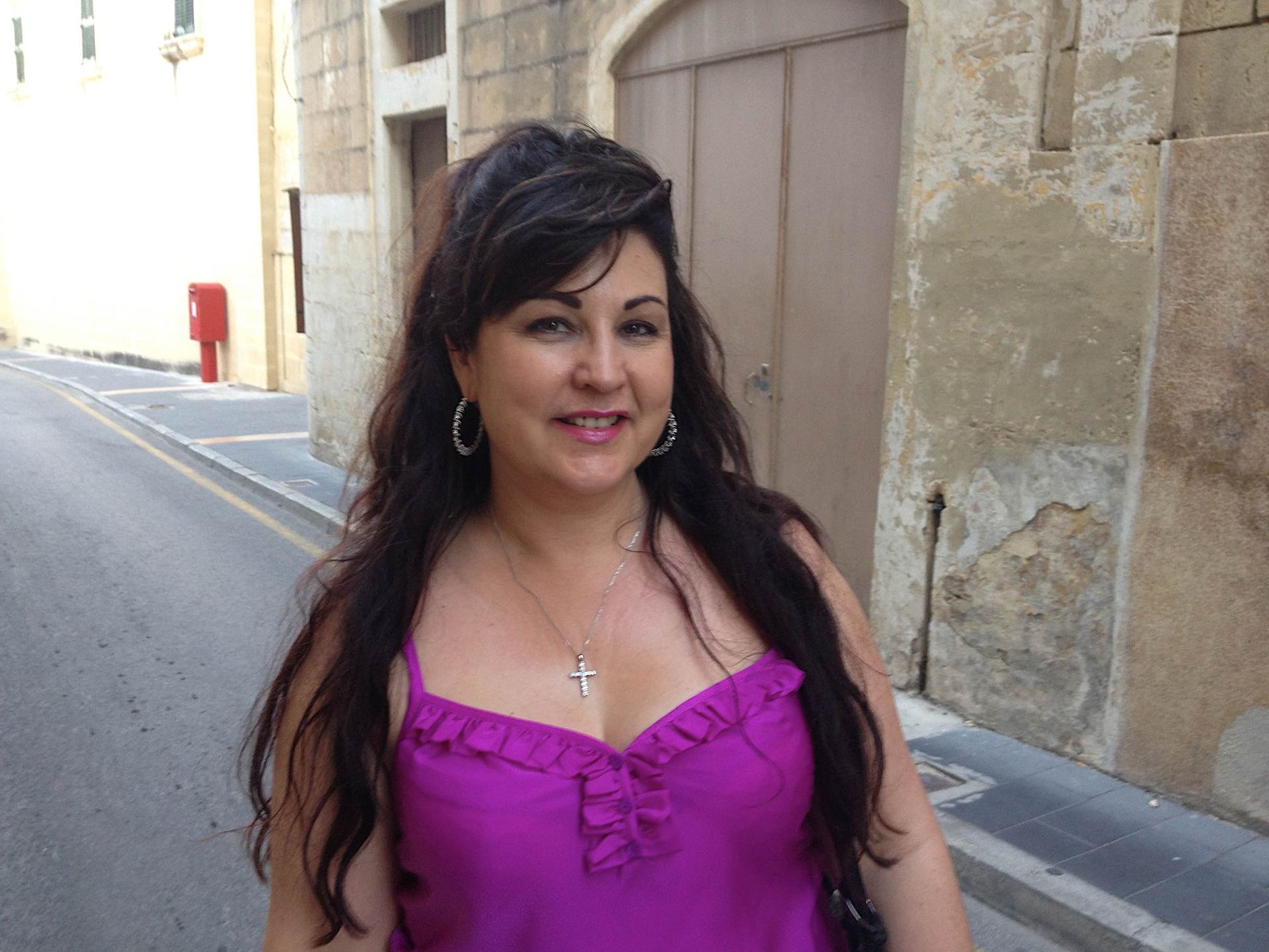Vivian Mizzi in a pink top smiles outdoors. Dark hair, silver necklace, stone buildingbbackdrop.