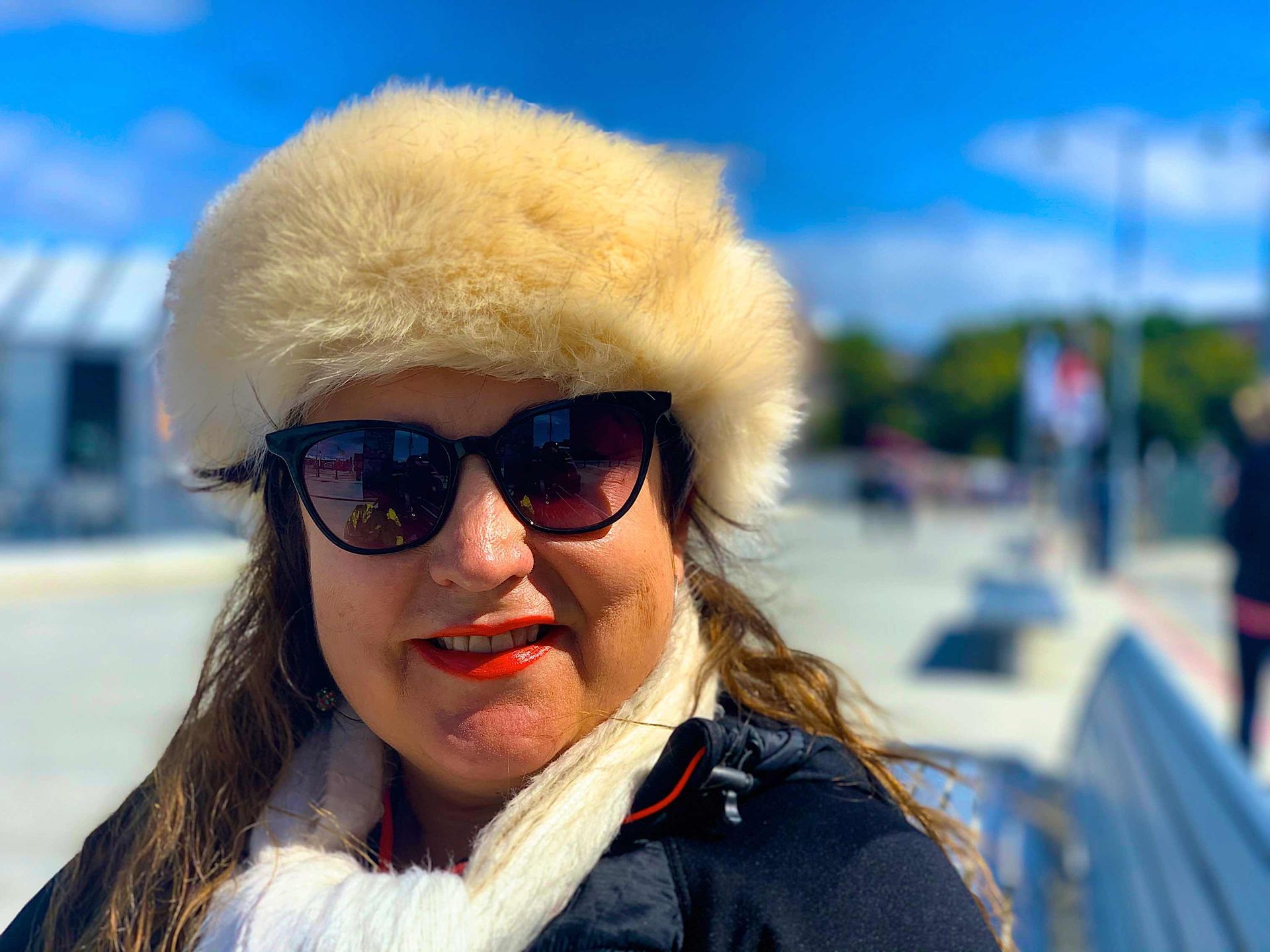 Vivian Mizzi with sunglasses and fur hat outdoors, smiling with red lipstick, blue sky in background.