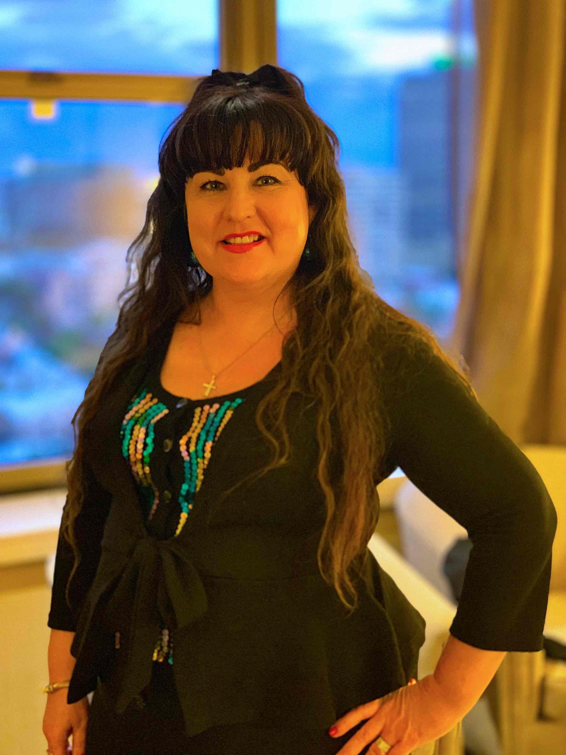 Vivian Mizzi with long dark hair, smiling, wearing a black cardigan and turquoise top, in front of a window.