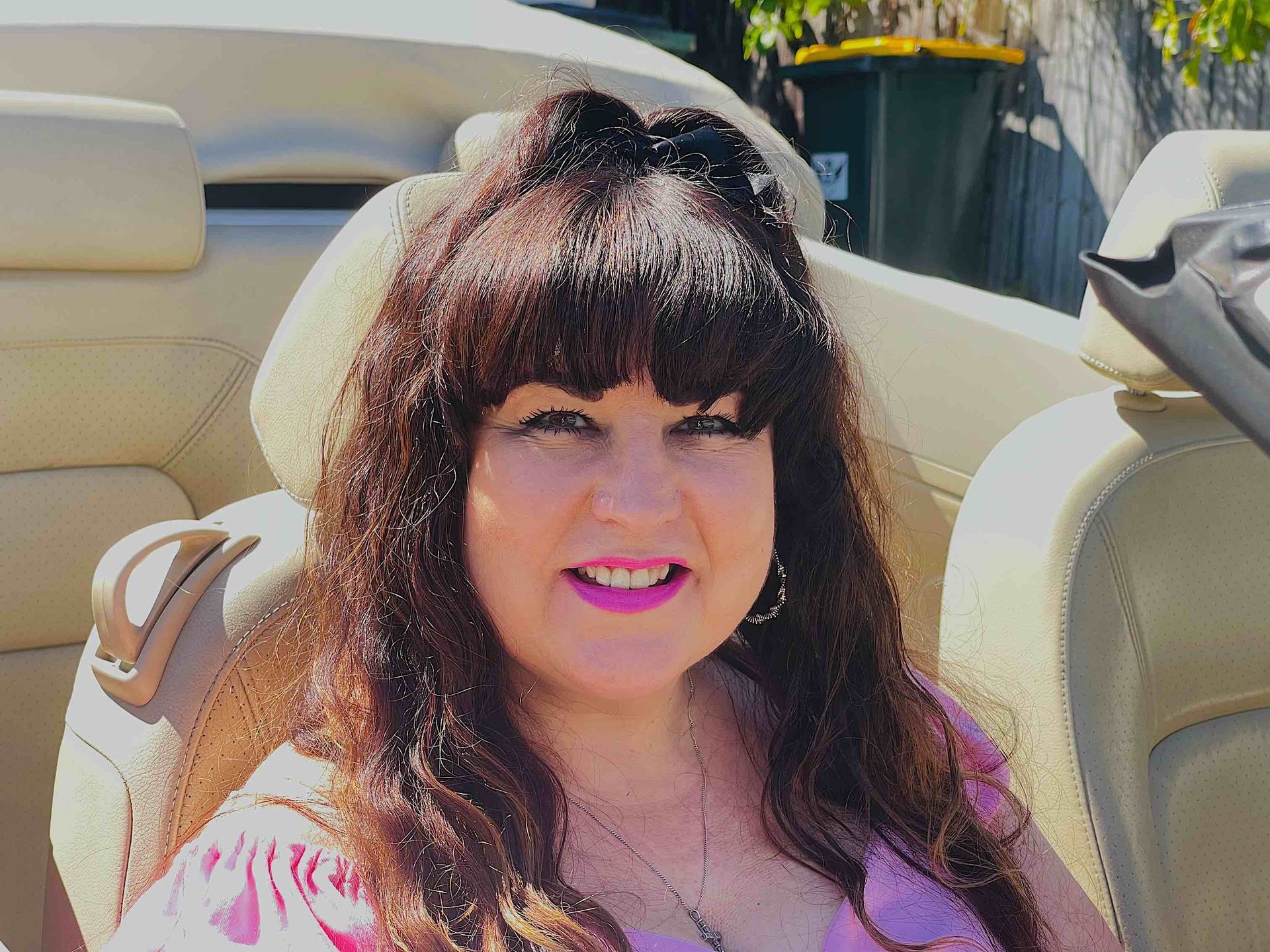 Vivian Mizzi with long brown hair in a convertible car, wearing pink, smiling at the camera.