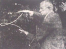 Man in coat, holding a rope loop outdoors, gesturing towards the loop, possibly dowsing.