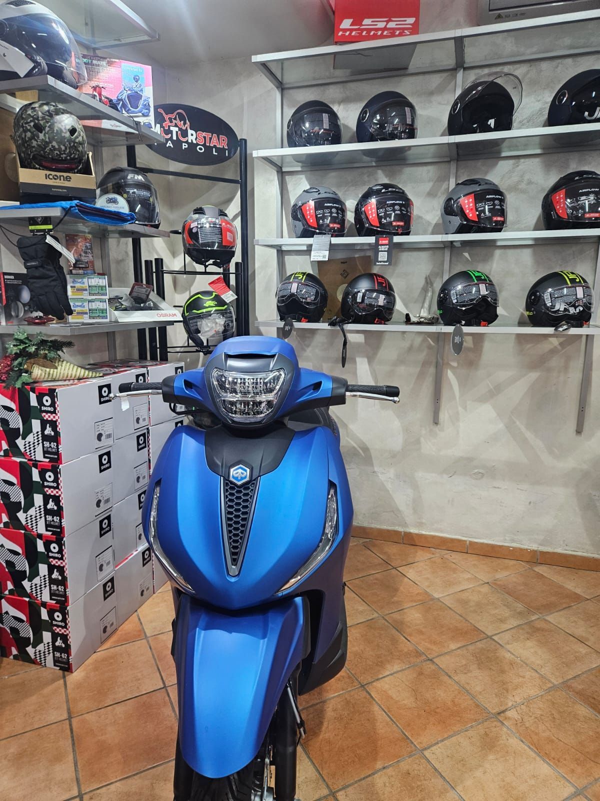 A blue scooter is parked in a room filled with helmets.