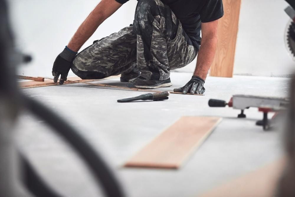 A man is kneeling down on the floor cutting a piece of wood.