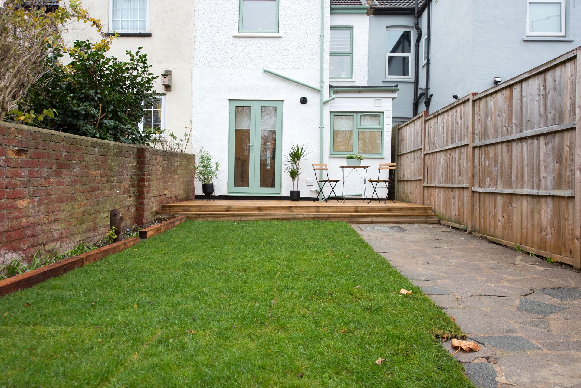 Backyard with wooden deck, green grass, and a brick wall. A light gray house is in the background.