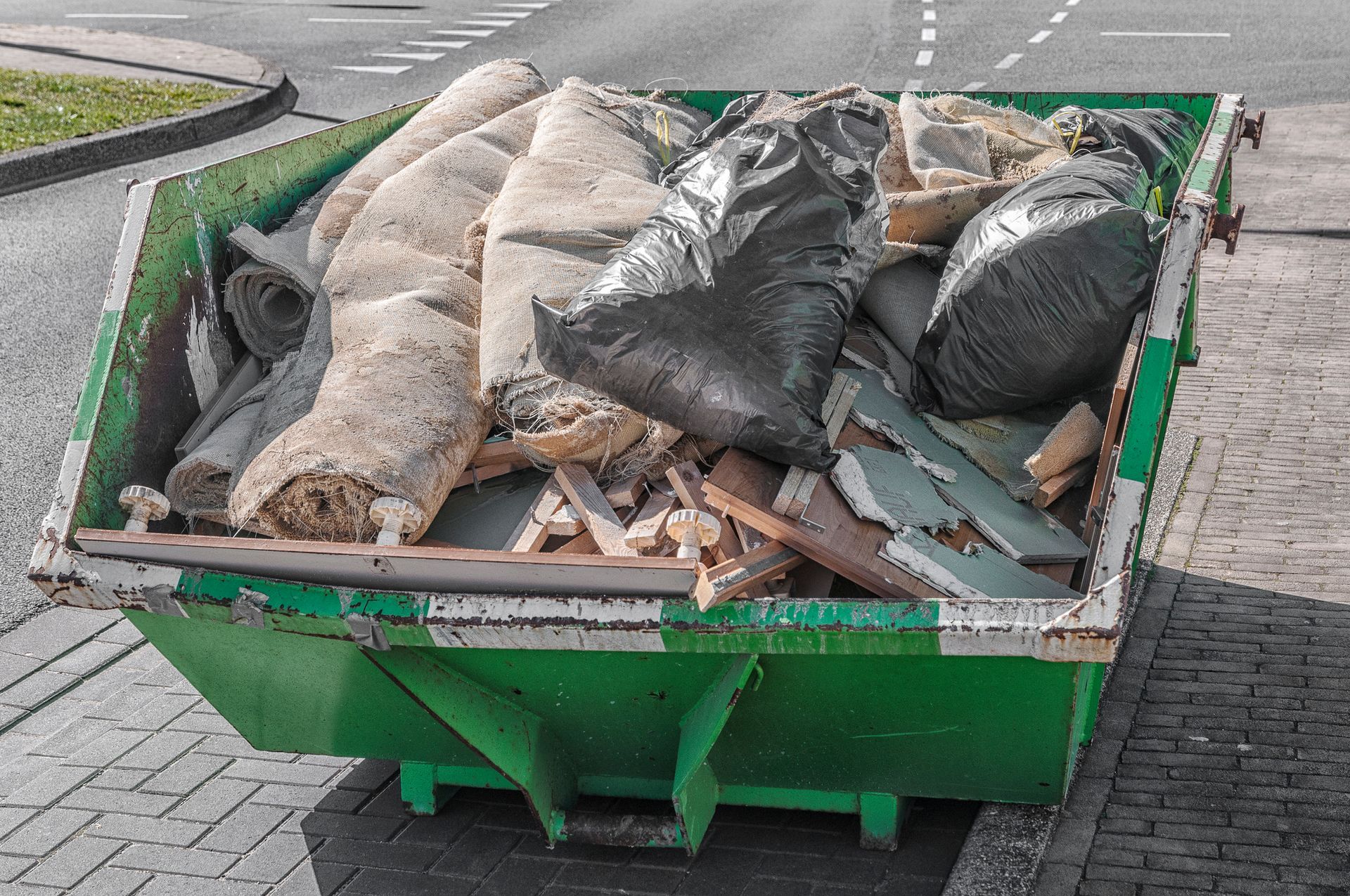 A large green steel waste container on the street full of waste and black plastic bags.