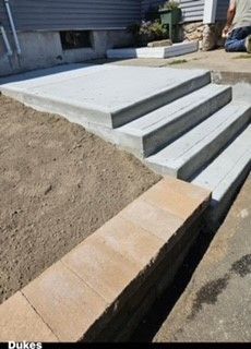 Concrete steps and landing newly built next to a tan brick retaining wall; dirt on one side, sidewalk on the other.