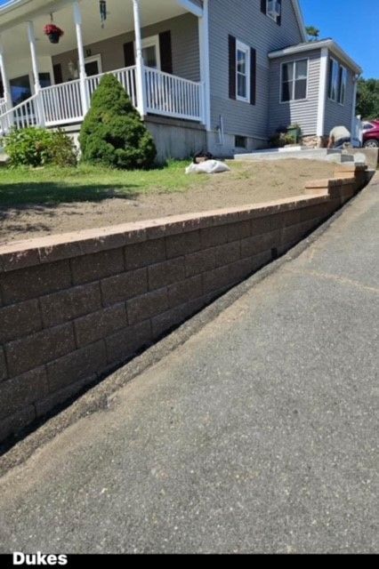 Retaining wall made of brown bricks beside a paved driveway, fronting a light gray house with a white porch.