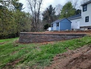 Retaining wall of dark blocks holds back soil near a blue shed and house on a grassy hill.