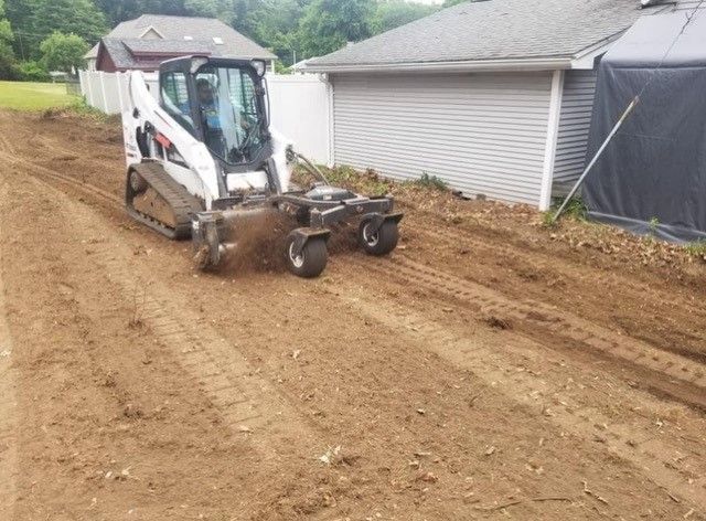 Bobcat skid-steer with a milling attachment, grinding a dirt surface outdoors near a building and fence.