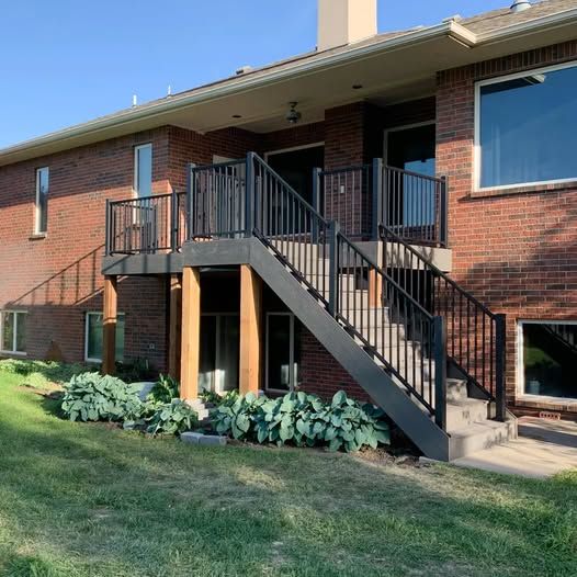 A brick house with a raised wooden deck featuring black railings and stairs leading down to a green lawn.