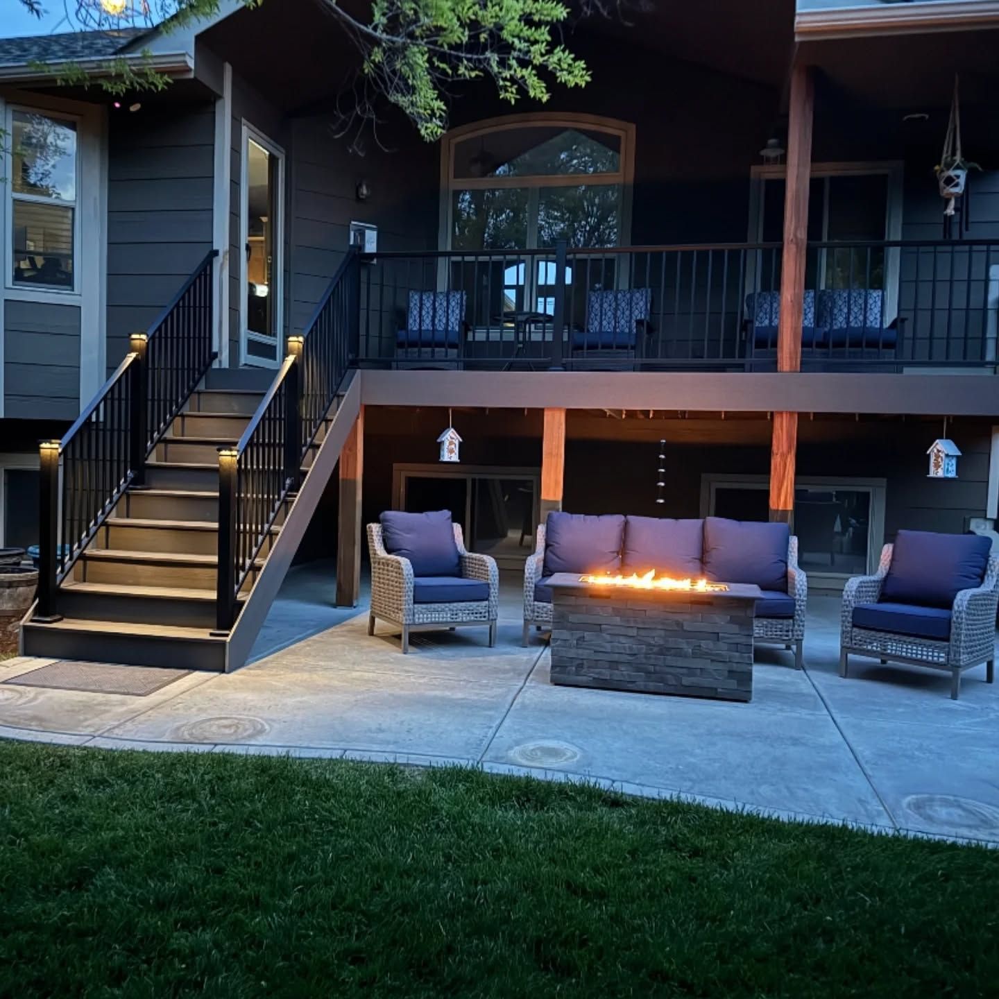 A patio with a lit fire pit and wicker furniture, situated beneath a wooden deck with stairs leading up to the house.