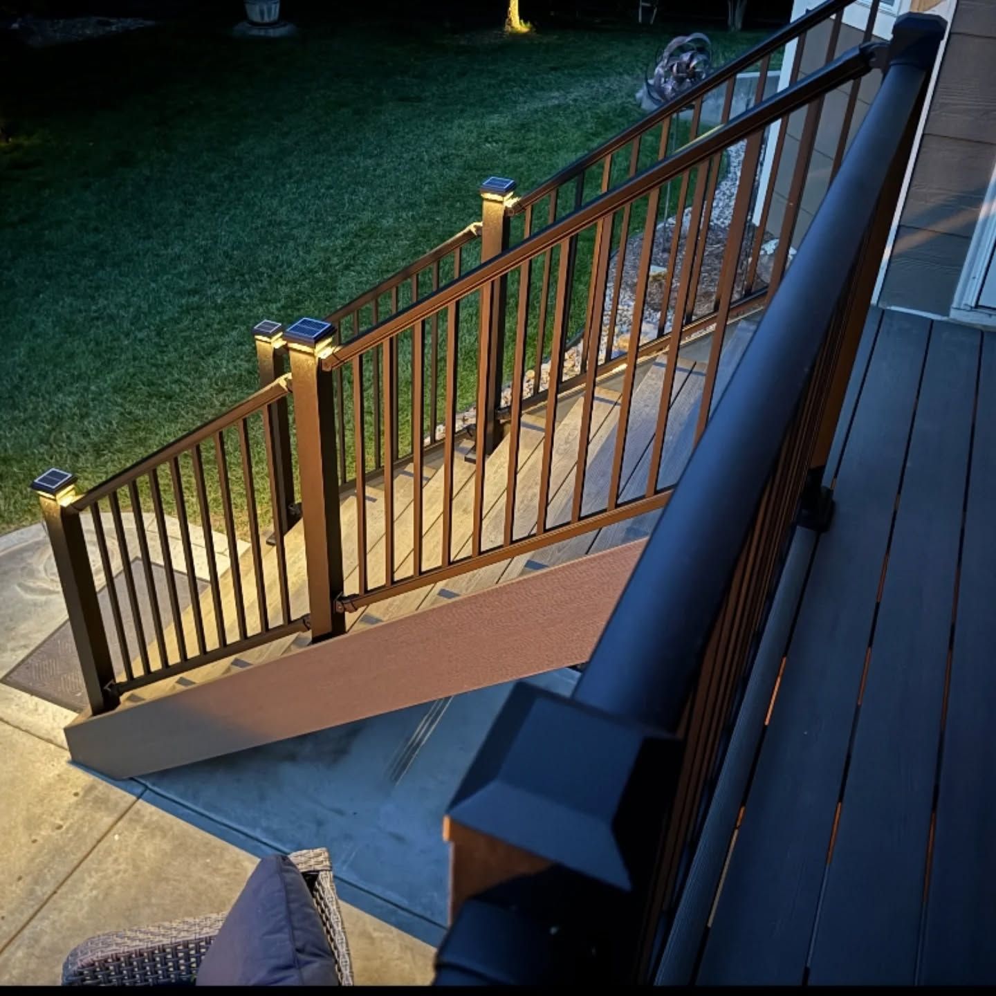 Dark metal railing and solar-powered post lights line a wooden staircase leading from a deck to a concrete patio at dusk.