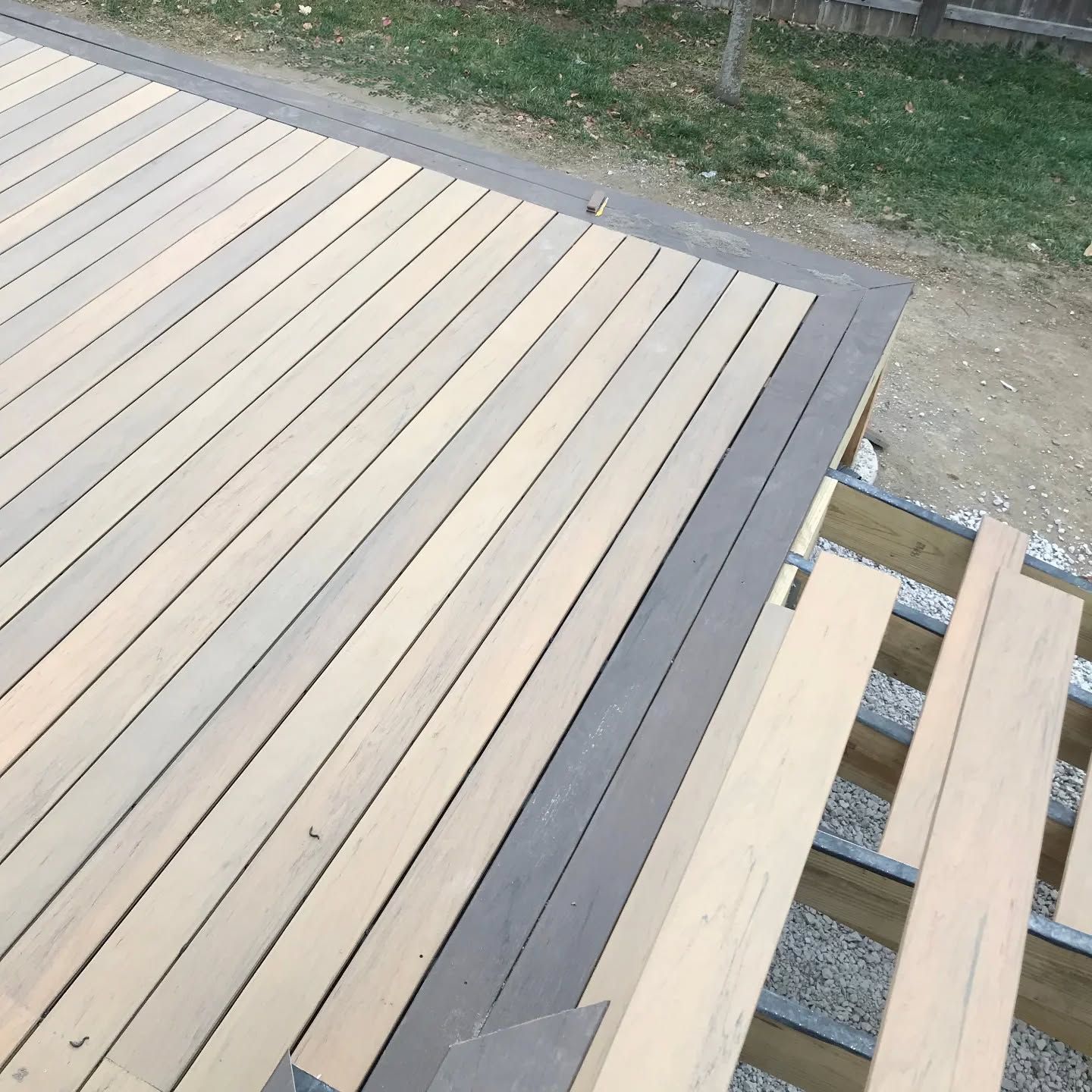 Construction of a deck showing rows of light-colored wood planks bordered by a dark gray trim piece on a metal frame.