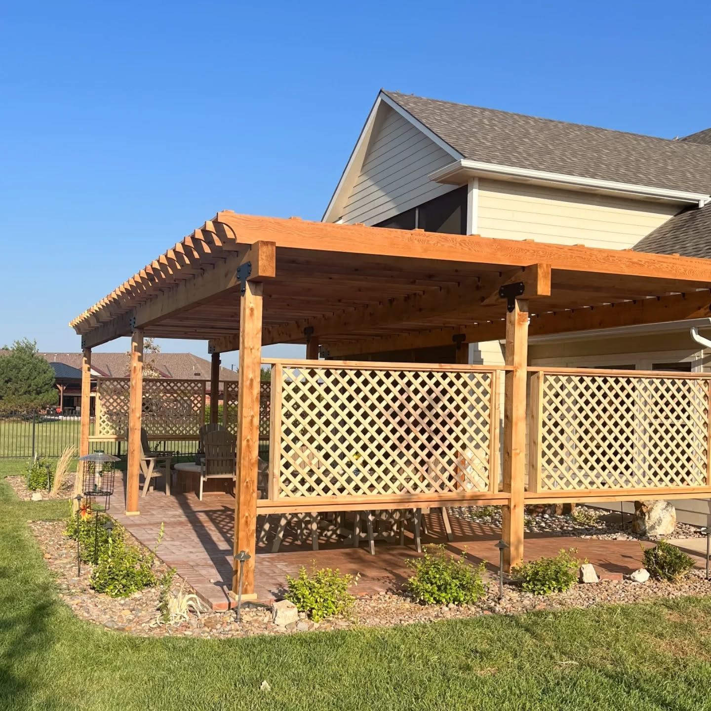 A wooden pergola with a lattice privacy screen sits on a patio next to a house with tan siding.