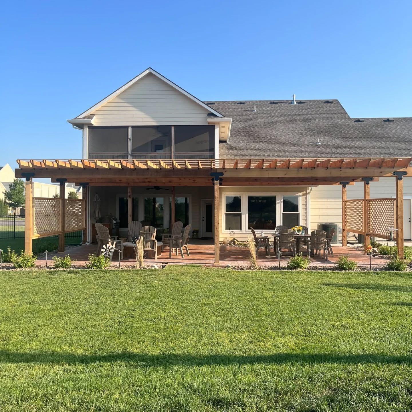 A wooden pergola covers a patio with patio furniture behind a house, with a lawn in the foreground under a clear sky.