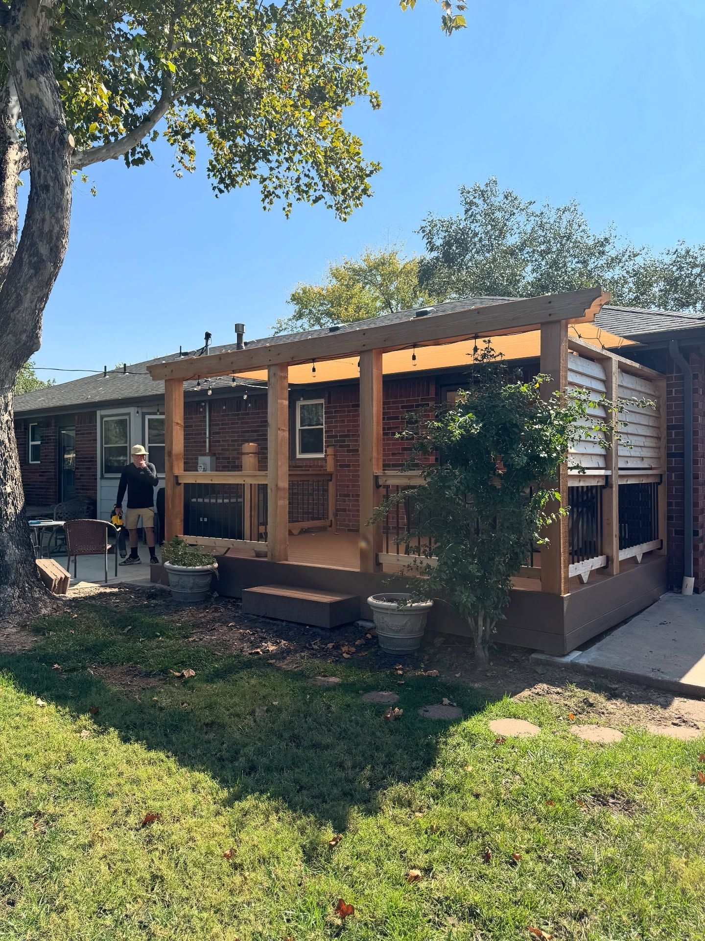 A wooden patio addition with a roof and lattice siding attached to the back of a brick house in a sunny backyard.